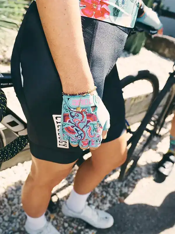 Woman wearing colorful floral cycling gloves and black shorts, standing beside a bike outdoors