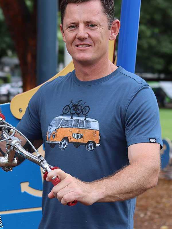 Man wearing cycling-themed graphic tee with van and bikes, holding bike handlebars outdoors