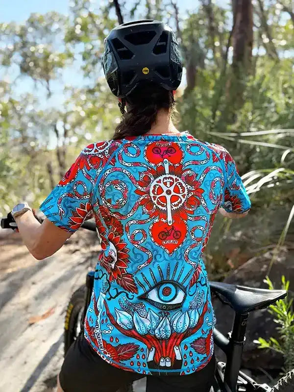 Woman cycling in vibrant blue and red patterned jersey with eye and heart design, forest trail backdrop
