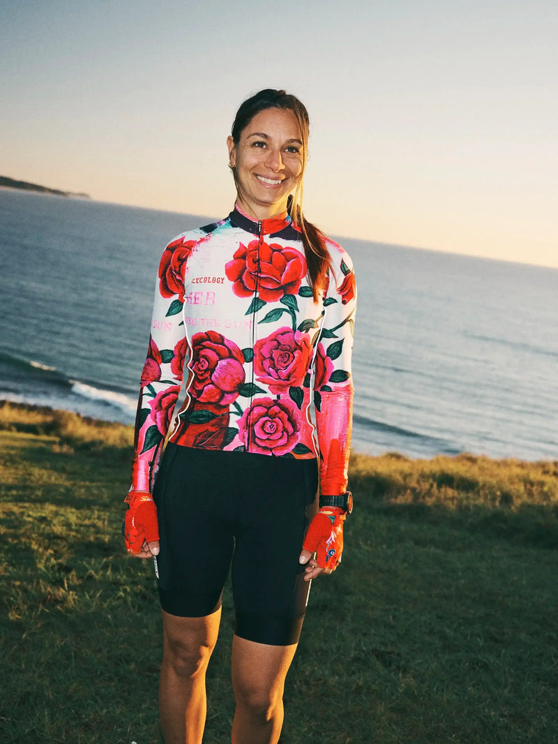 Woman in floral cycling jersey and black shorts, smiling by ocean at sunset