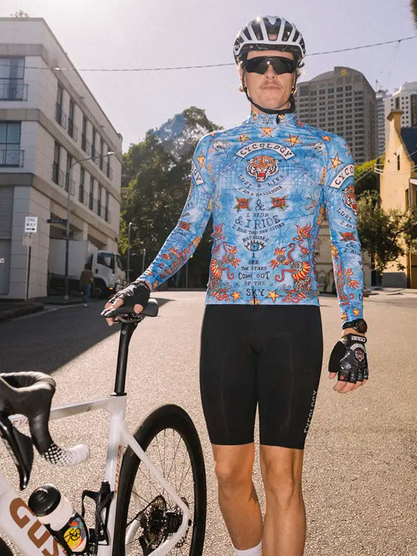 Cyclist in a Blue men's long sleeve cycling jersey with colorful graphics  and black bib shorts standing next to a bicycle on a city street.