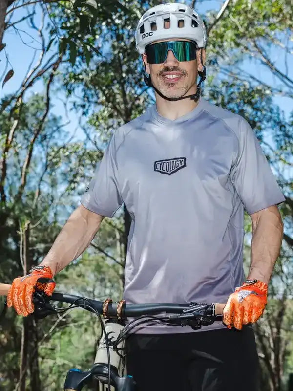 Man wearing grey cycling jersey with Cycology logo, orange gloves, and POC helmet on mountain bike trail