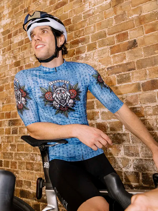 Person wearing a blue cycling jersey with floral wolf designs sitting on a bike against a brick wall.