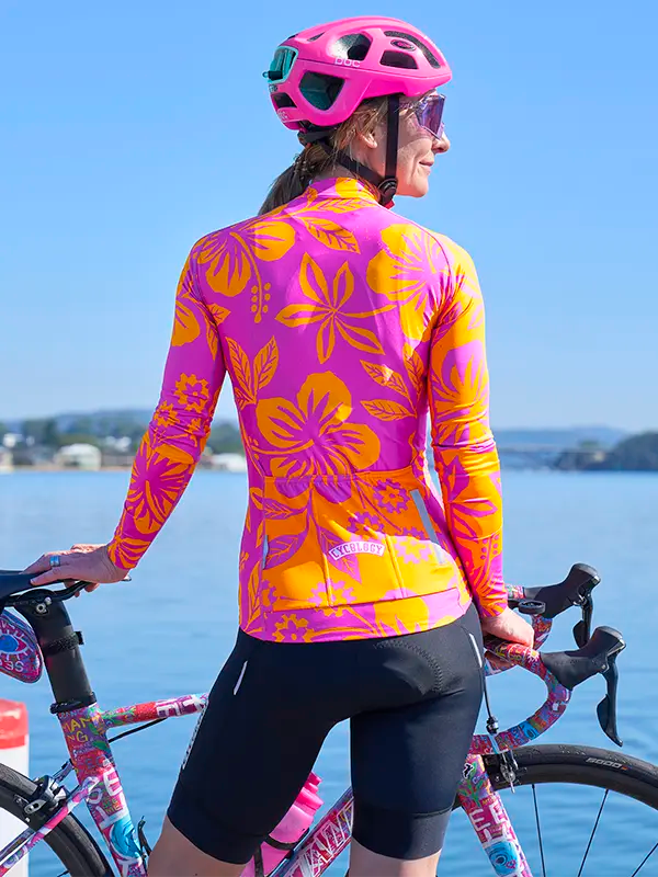 Person wearing a colorful Women's Summer Long Sleeve Jersey with a pink helmet and sunglasses, standing next to a bicycle by a body of water.