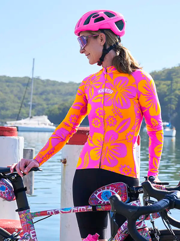 Person wearing a colorful Women's Summer Long Sleeve Jersey with a pink helmet and sunglasses, standing next to a bicycle by a waterfront.