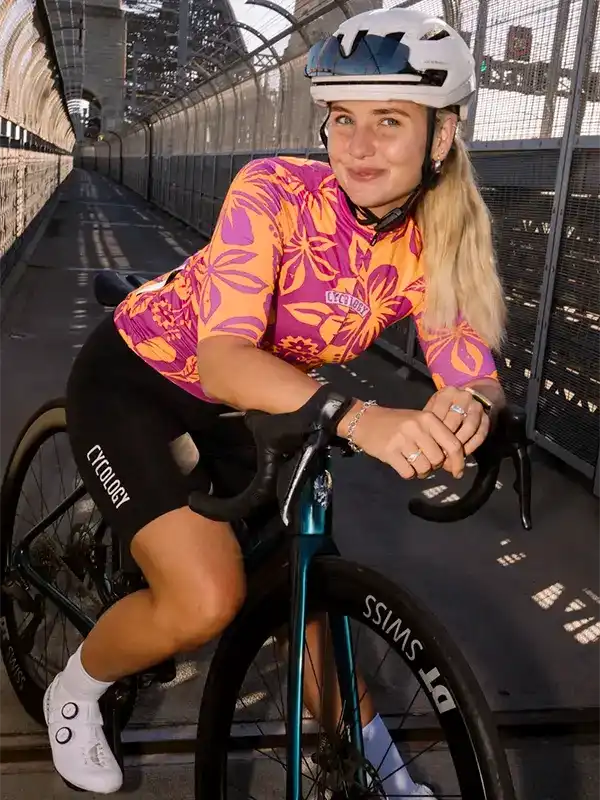 Woman in vibrant floral cycling jersey and black shorts riding on a bridge, wearing helmet and gloves.