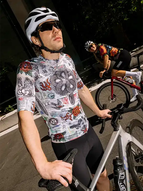 Man in tattoo-style cycling jersey and helmet, riding road bike with woman in floral kit nearby