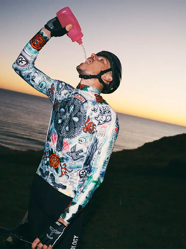 Person wearing a colorful cycling jersey with a helmet, pouring a pink bottle against a sunset sky.