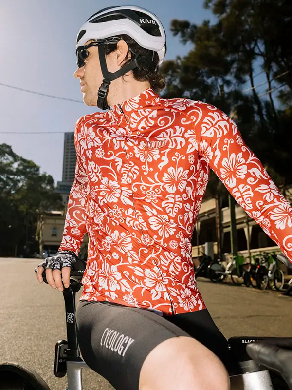 Cyclist wearing a red and white floral cycling jersey with a helmet and gloves, standing on a street.