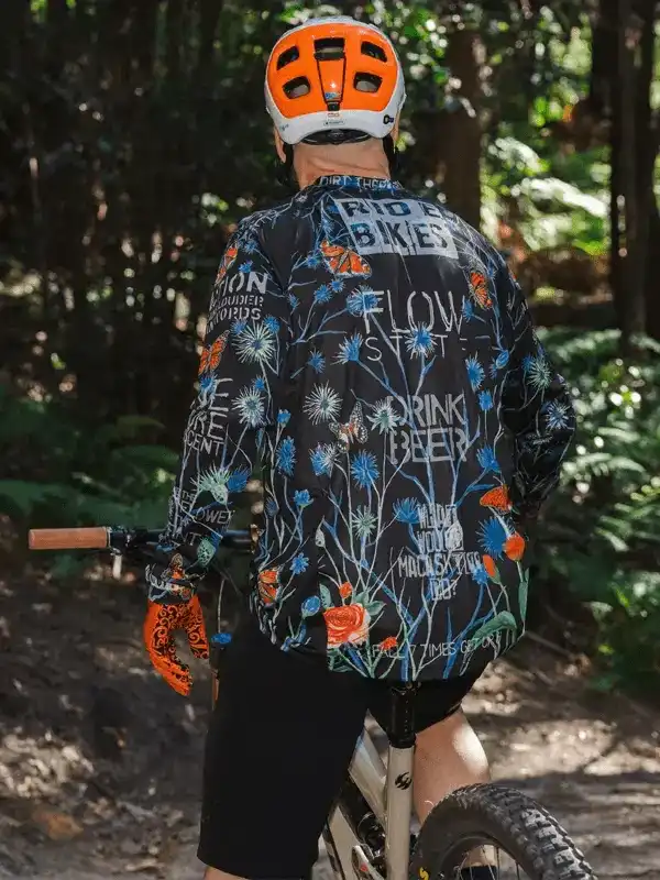 Cyclist wearing floral print long sleeve jersey with 'Ride Bikes' text, orange helmet, on forest trail