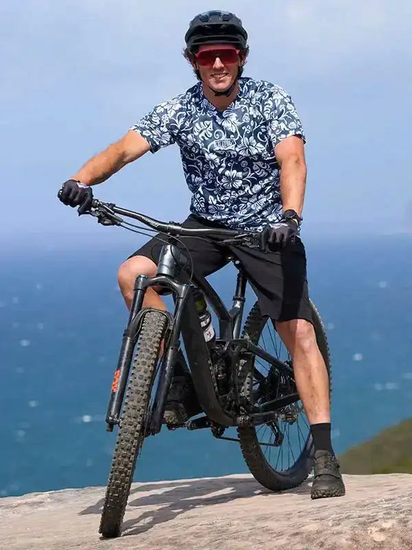 Man riding mountain bike in floral cycling jersey, helmet, and sunglasses on coastal cliff